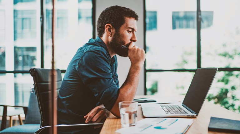 Male sitting at and leaning on desk looking thoughtfully at his laptop with hand at his mouth